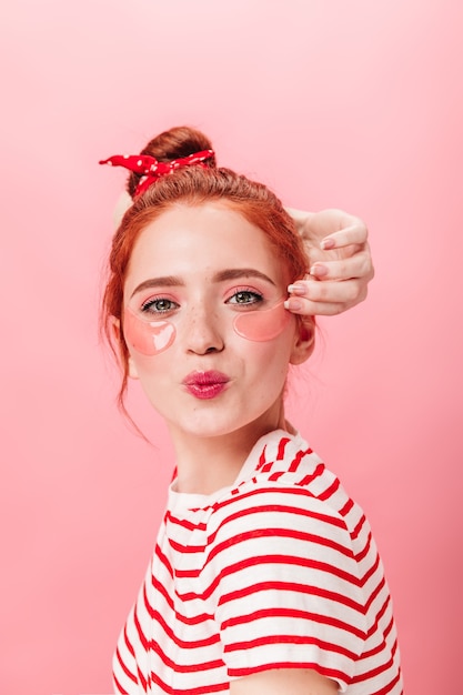 Ragazza sicura dello zenzero con bende sull'occhio che guarda l'obbiettivo. Studio shot di gioiosa donna caucasica in posa con baciare l'espressione del viso su sfondo rosa.