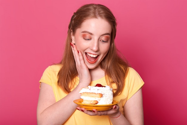 ragazza soddisfatta soddisfatta con i capelli castano chiaro, tiene un enorme pezzo di gustosa torta, tiene la bocca aperta, piena di divertimento, vestita con una maglietta gialla casual