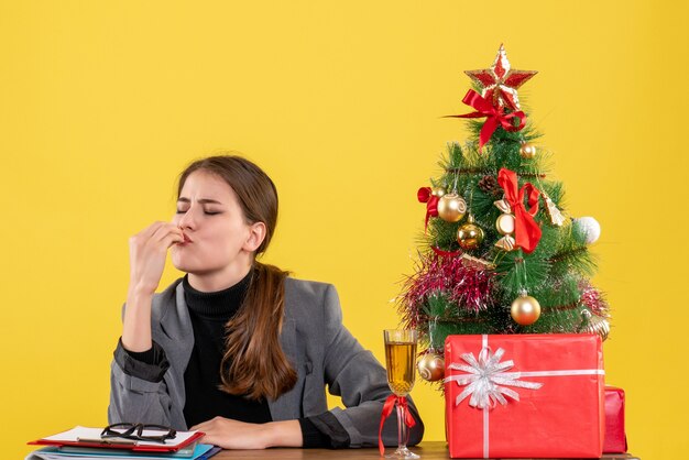 Ragazza soddisfatta vista frontale che si siede allo scrittorio che fa il segno di bacio del capo s vicino all'albero di Natale e al cocktail dei regali