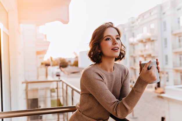 Ragazza sognante dagli occhi scuri che beve il tè al balcone. Foto del modello femminile ben vestito caucasico che tiene la tazza di caffè.