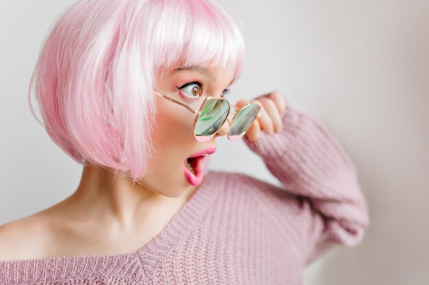 Ragazza sorpresa con i grandi occhi che distoglie lo sguardo durante il servizio fotografico. Foto del primo piano della giovane donna sensuale in peruke e occhiali da sole rosa.