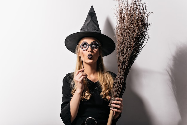 Ragazza sorpresa con labbra nere in posa al carnevale di halloween. Splendida signora dai capelli lunghi in costume da strega in piedi sul muro bianco.