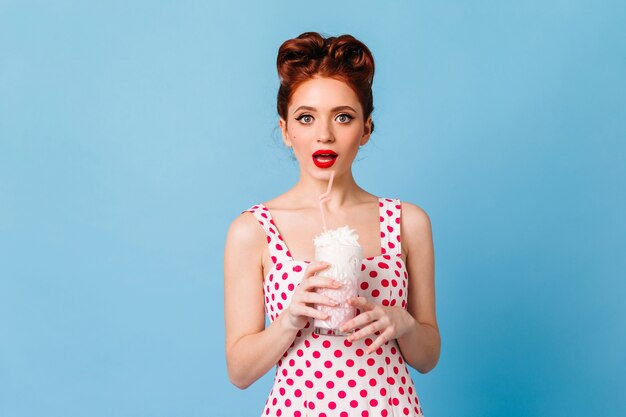 Ragazza sorpresa con le labbra rosse che beve frappè. Studio shot di emotiva giovane donna in abito a pois in piedi sullo spazio blu.