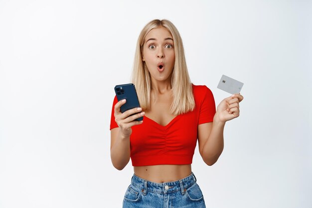 Ragazza sorpresa con telefono cellulare e carta di credito che sembra impressionata dalla reazione della fotocamera allo shopping online in vendita nell'applicazione su sfondo bianco dello smartphone