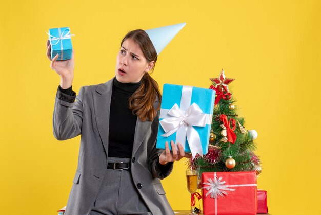 Ragazza sorpresa di vista frontale con la protezione del partito che tiene i regali di natale vicino all'albero di Natale e al cocktail dei regali