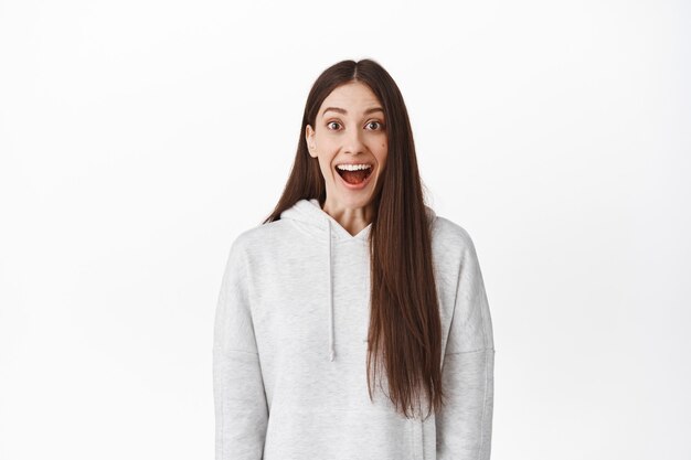 Ragazza sorpresa ed elettrizzata sorridente, in piedi in soggezione con la bocca aperta e guardando davanti all'evento affascinato, mozzafiato ed emozionante, in posa contro il muro bianco