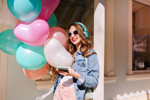 Ragazza sorridente allegra in occhiali da sole alla moda andando all'evento e ascoltando la musica preferita in cuffia. Adorabile giovane donna che indossa giacca di jeans retrò che trasportano palloncini colorati alla festa di compleanno.