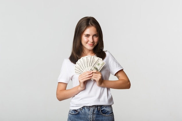 Ragazza sorridente avida che sembra astuta e che tiene soldi, bianca in piedi.