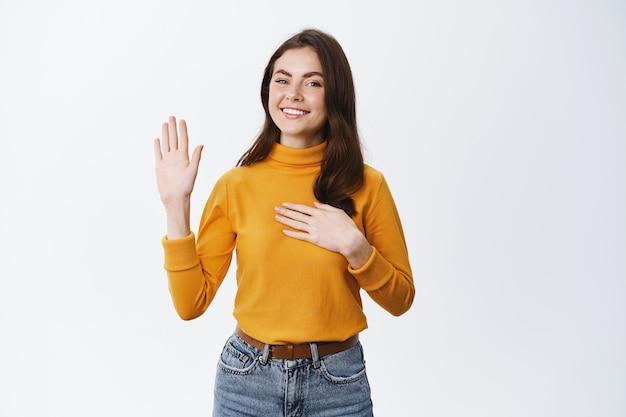 Ragazza sorridente che fa una promessa, alza una mano e ne mette un'altra sul cuore, giura di essere la verità e sii onesta, in piedi contro il muro bianco