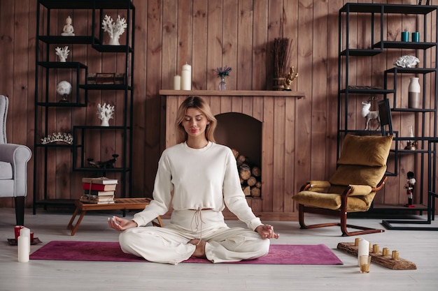 Ragazza sorridente che pratica yoga che si siede nella posa del loto, meditando nell'interno domestico accogliente. Formazione femminile per il benessere.