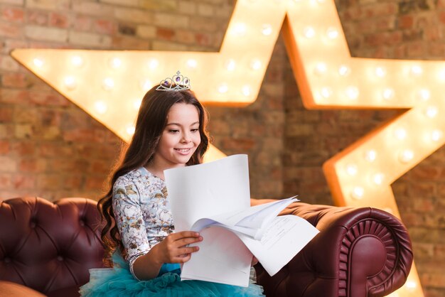 Ragazza sorridente che si siede sugli scritti della lettura del sofà contro la stella d&#39;ardore nella priorità bassa