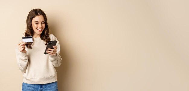 Ragazza sorridente che utilizza l'app mobile smartphone shopping e carta di credito in piedi su sfondo beige ord