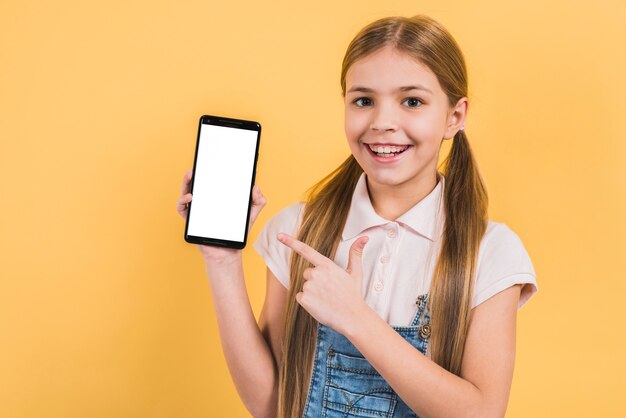 Ragazza sorridente con capelli biondi lunghi che indica la sua barretta al telefono mobile in bianco dello schermo bianco contro fondo giallo