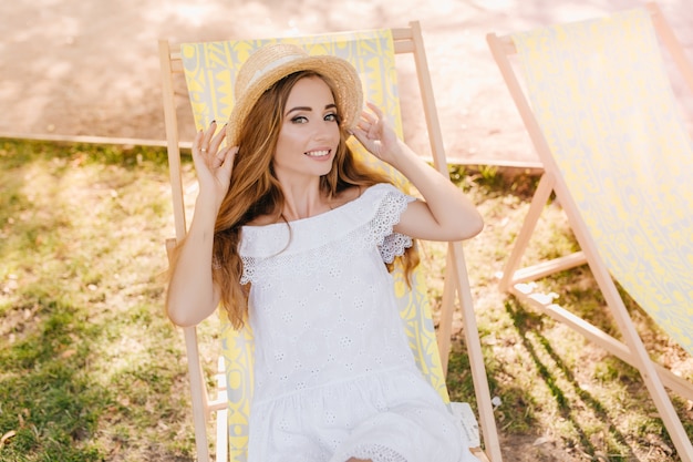 Ragazza sorridente con trucco alla moda rilassante in chaise-longue di buon umore, con cappello di paglia. Foto all'aperto di felice giovane donna in abito bianco godendo il fine settimana in giardino.