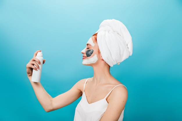 Ragazza sorridente con un asciugamano sulla testa tenendo la bottiglia di cosmetici. Studio shot di ridere donna con maschera facciale in piedi su sfondo blu.