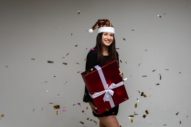 Ragazza sorridente con un grande regalo festivo che punta a mano su uno spazio vuoto con coriandoli festivi su sfondo grigio