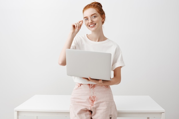 Ragazza sorridente della testarossa che per mezzo del computer portatile e che osserva