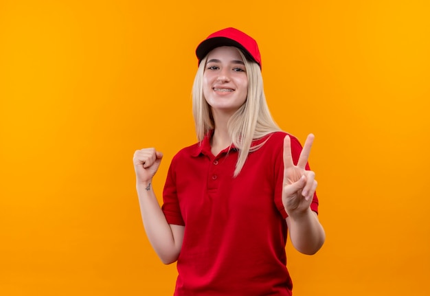 Ragazza sorridente di consegna che indossa la maglietta e il cappuccio rossi in parentesi graffa dentale che mostra i gesti di sì e di pace su fondo arancio isolato
