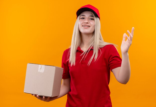 Ragazza sorridente di consegna che indossa la maglietta rossa e il berretto in scatola della tenuta del tutore dentale e mano sollevata su fondo arancio isolato