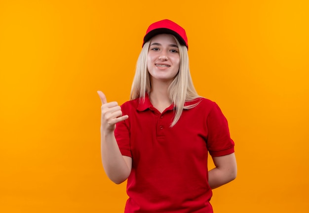 Ragazza sorridente di consegna che indossa la maglietta rossa e il cappuccio in parentesi graffa dentale che mostra il gesto del telefono su fondo arancio isolato