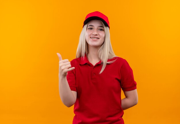 Ragazza sorridente di consegna che indossa la maglietta rossa e il cappuccio in parentesi graffa dentale che mostra il gesto del telefono su fondo arancio isolato
