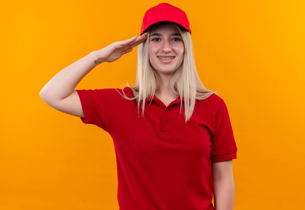 Ragazza sorridente di consegna che porta la maglietta e il cappuccio rossi in parentesi graffa dentale che mostra gesto di saluto su fondo arancio isolato