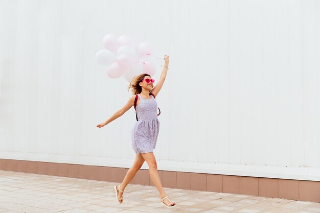 Ragazza sorridente emozionante in occhiali da sole rosa in esecuzione con palloncini, indossando abiti e sandali