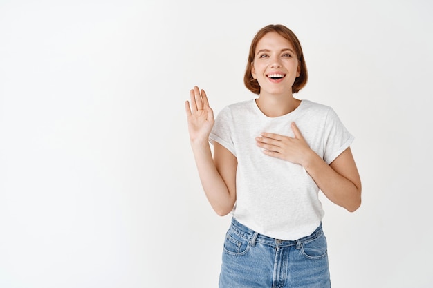 Ragazza sorridente felice che alza il braccio e mette la mano sul cuore, essendo onesta, dicendo la verità, giura di essere sincera, in piedi contro il muro bianco