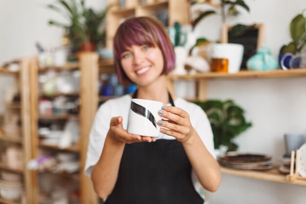 Ragazza sorridente in grembiule nero e maglietta bianca che tiene in mano una tazza fatta a mano che la mostra sulla fotocamera allo studio di ceramiche