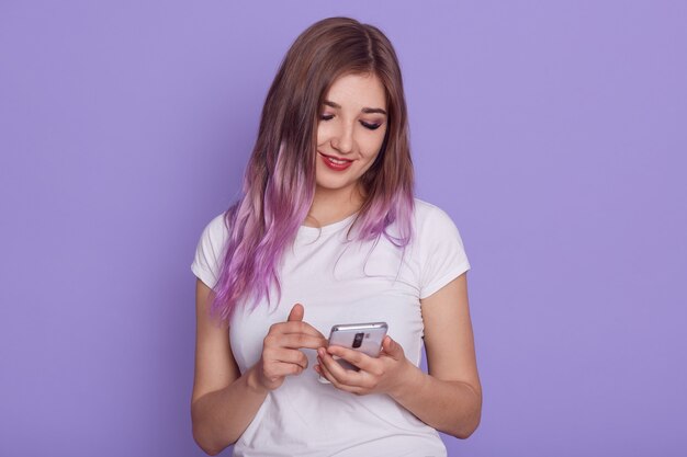 Ragazza sorridente in maglietta casual bianca utilizzando il telefono cellulare moderno, tenendo il dispositivo nelle mani, guardando il display con espressione positiva, in posa isolato sopra la parete viola.