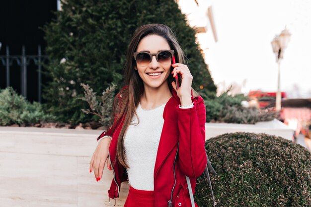 Ragazza sorridente in occhiali da sole scuri parlando al telefono mentre posa vicino al cespuglio verde