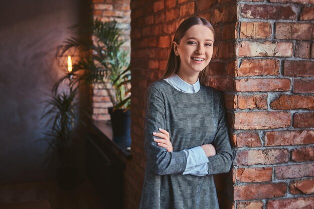 Ragazza sorridente vestita con un elegante abito grigio in piedi con le braccia incrociate mentre si appoggia al muro di mattoni, guardando una telecamera.