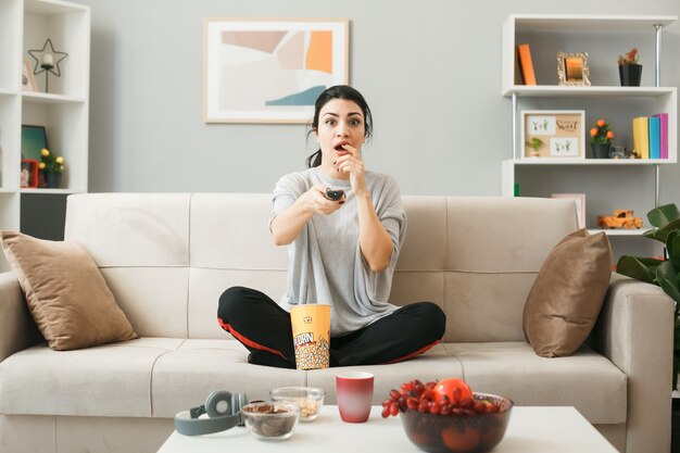 Ragazza spaventata con secchio di popcorn che tiene il telecomando della tv, seduta sul divano dietro il tavolino da caffè nel soggiorno