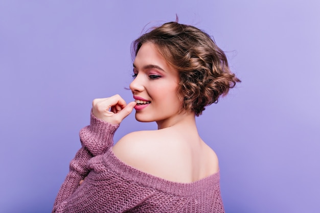 Ragazza spaventata in sciarpa lavorata a maglia bianca che copre il viso con le mani. Ritratto dell'interno della giovane donna europea emotiva in cappello isolato sulla parete viola.