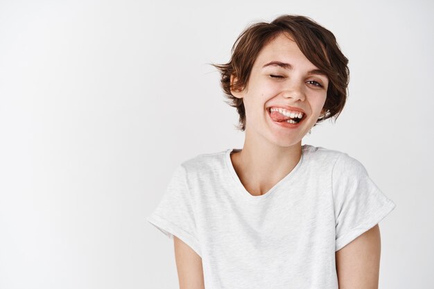 Ragazza spensierata con i capelli corti, strizzando l'occhio e mostrando la lingua con una faccia sciocca e felice, in piedi felice in maglietta contro il muro bianco