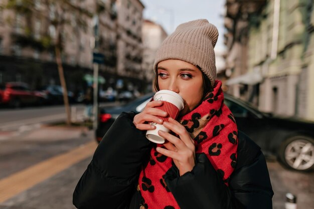 Ragazza spensierata con un ottimo trucco che beve tè su una strada soleggiata nel centro della città Foto di una piacevole donna bruna con una giacca scura che si gode il caffè