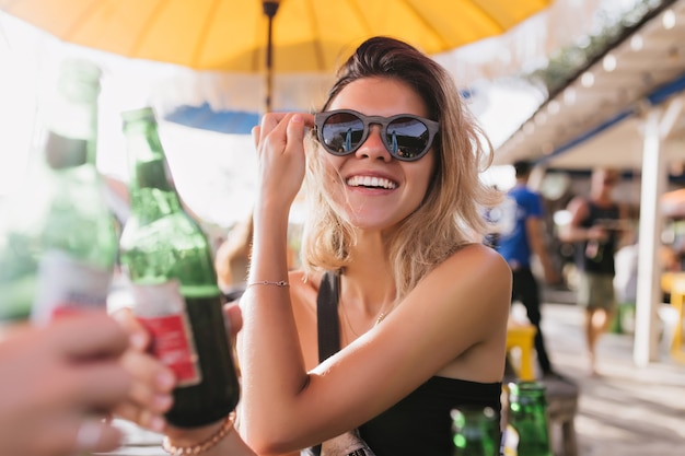 Ragazza spensierata di risata che beve birra nella caffetteria estiva. Bella signora abbronzata in occhiali da sole in posa con piacere in una giornata calda.