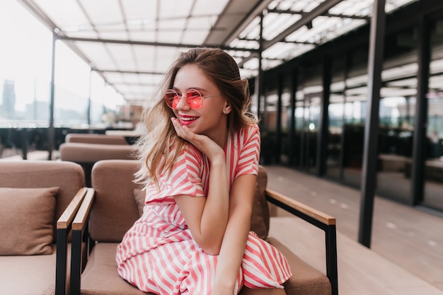 Ragazza spensierata di risata che si siede nella caffetteria e guardandosi intorno. Magnifica donna bionda in abito a righe vintage che esprime emozioni positive.