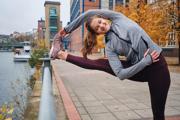 Ragazza sportiva abbastanza sorridente che si allunga felicemente durante l'allenamento sulla strada vuota della città