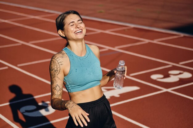 Ragazza sportiva allegra in abiti sportivi felicemente acqua potabile dopo l'allenamento sullo stadio cittadino