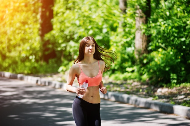 Ragazza sportiva fitness in abbigliamento sportivo correre su strada nella foresta con acqua in bottiglia a portata di mano sport all'aria aperta in stile urbano
