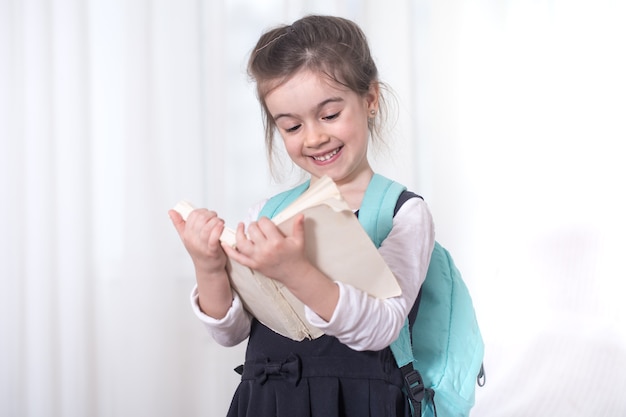 Ragazza-studentessa di scuola elementare con uno zaino sulle spalle leggendo un libro su uno sfondo chiaro. Il concetto di istruzione e scuola primaria