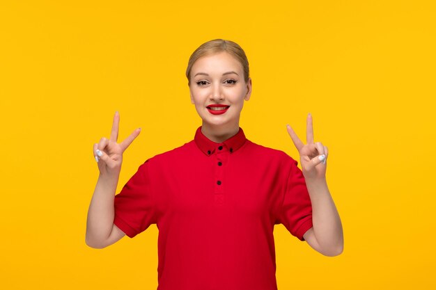 Ragazza sveglia di giorno della camicia rossa che mostra il segno di pace in una camicia rossa su una priorità bassa gialla