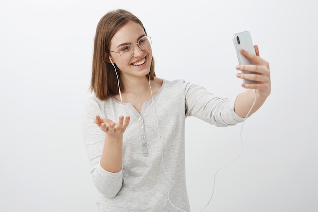 Ragazza talkign con un amico tramite messaggio video gesticolando con la mano durante una divertente conversazione che punta la fotocamera dello smartphone al viso che indossa gli auricolari e sorride allo schermo del dispositivo oltre il muro grigio