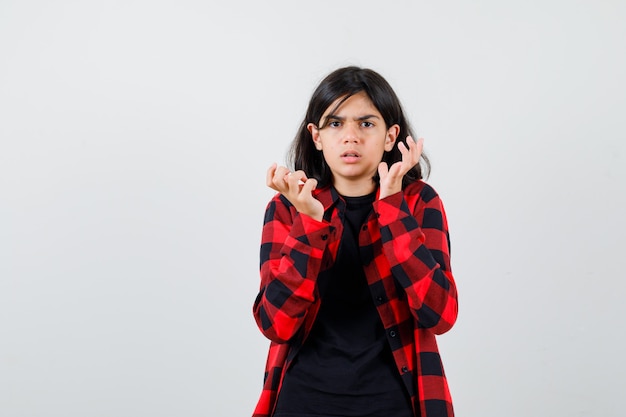 Ragazza teenager che alza le mani vicino al viso in maglietta, camicia a scacchi e sembra perplessa. vista frontale.