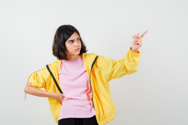 Ragazza teenager che indica in su con il dito, tenendo la mano sulla vita in tuta gialla, t-shirt e guardando pensieroso, vista frontale.