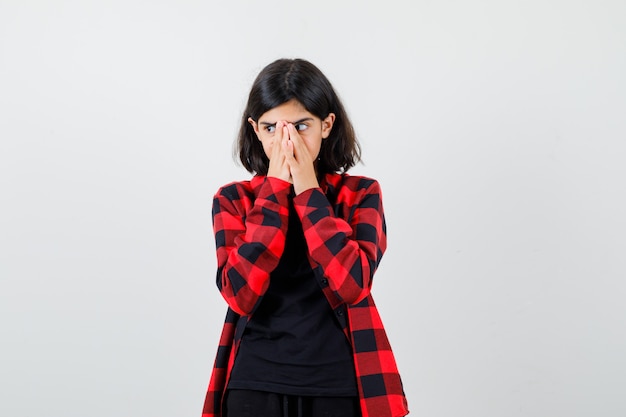 Ragazza teenager che si tiene per mano sulla bocca in t-shirt, camicia a scacchi e guardando premurosa, vista frontale.