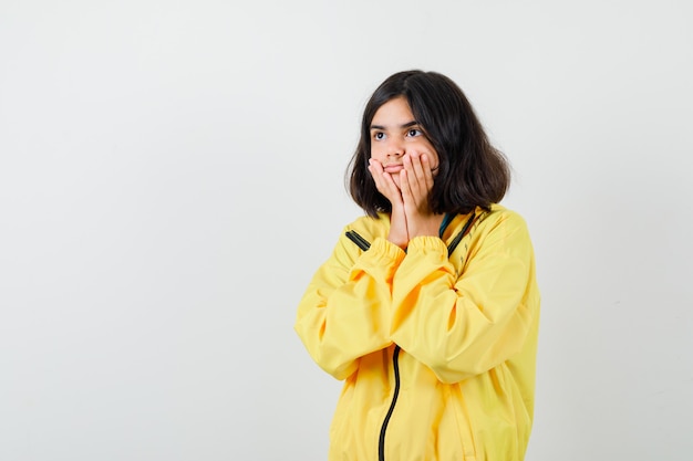 Ragazza teenager che si tiene per mano sulle guance, distogliendo lo sguardo in giacca gialla e guardando perplesso, vista frontale.