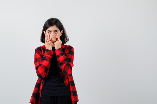 Ragazza teenager che tira giù le palpebre in maglietta, camicia a scacchi e sembra delusa, vista frontale.