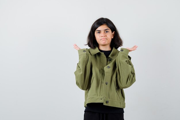 Ragazza teenager in giacca verde militare che mostra gesto impotente e sembra perplessa, vista frontale.